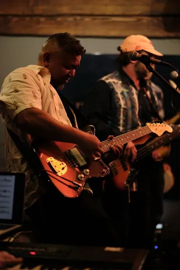The CJP Band playing live — guitarist and bassist on stage under warm lights